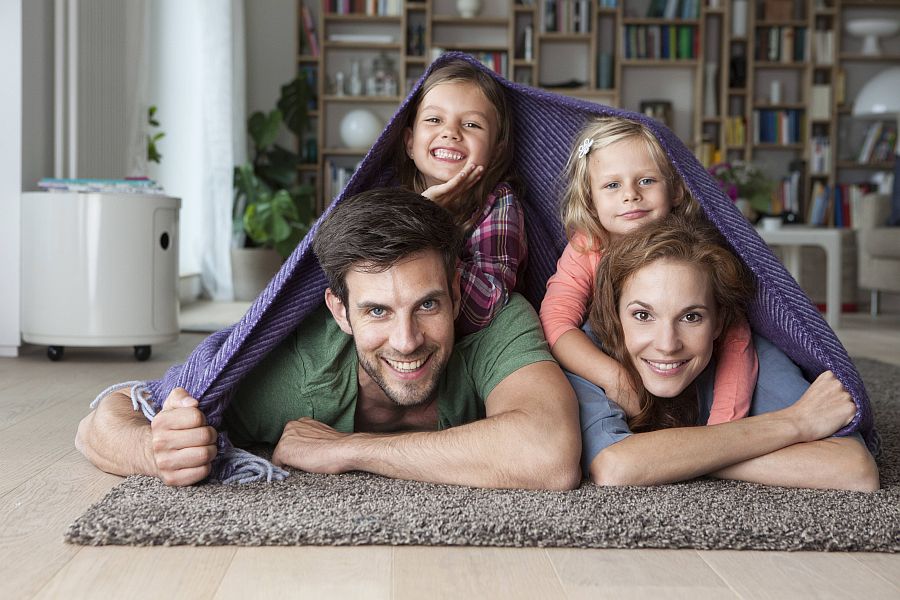 Vater, Mutter und zwei Kinder haben eine Decke wie ein Zelt über sich ausgebreitet und liegen auf dem Wohnzimmerteppich