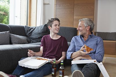 Vater und Teenager-Sohn sitzen auf dem Wohnzimmerboden, essen Pizza und schauen sich lachend an.