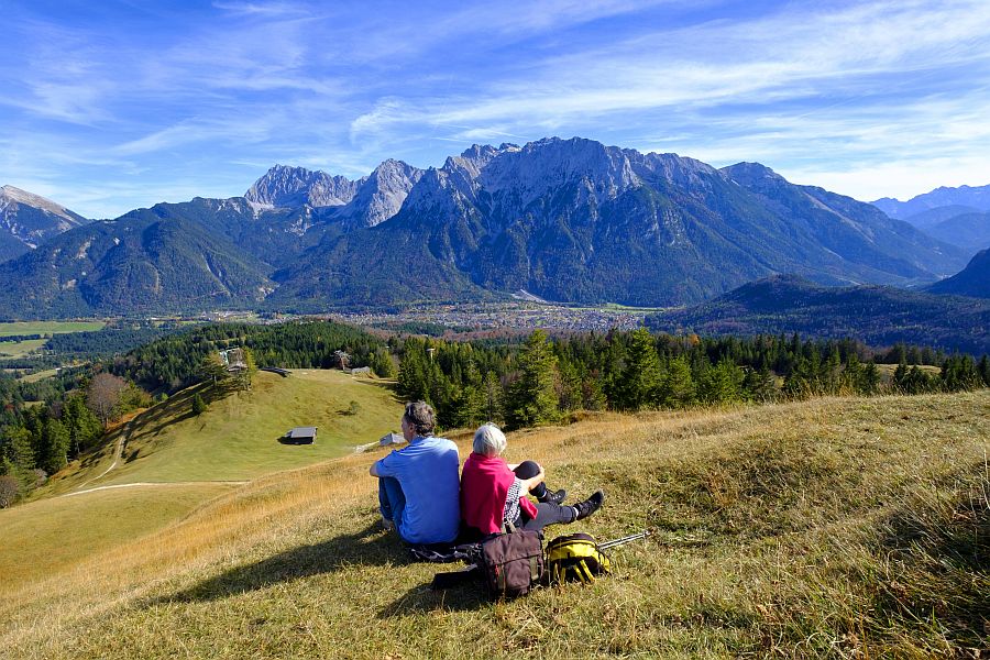 Älteres Paar sitzt auf einer sommerlichen Bergwiese und sieht hinab ins Tal, neben ihnen liegen ihre Rucksäcke.